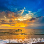 A fishing boat goes to the ocean in the evening. Bright colorful orange sun hidden in dense blue clouds over the Bali Sea. A white wave pours a foam to the sandy beach.